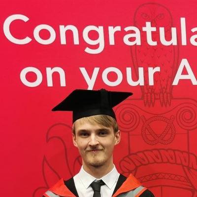 Fergus is wearing a cap and gown for graduation, smiling brightly into the camera. Behind him are the words "Congratulations on your Award" in white font on a red background.