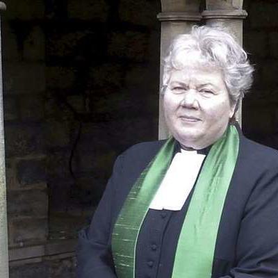 Reverend Elspeth McKay is standing in front of a stone building in traditional reverend attire, including a green stole. She is smiling into the camera with her hands held in front of her.