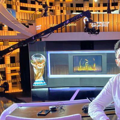 Saqib Uddin is sitting at a desk within a TV studio smiling brightly into a camera. Behind him is signage for the “World Cup Qatar 2022”.