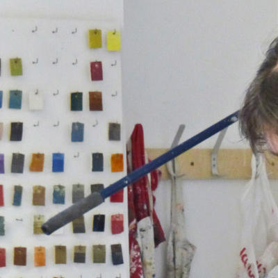 Sian is standing over a large bowl filled with clay. They are wearing a blue polo shirt which is covered by a white apron with various colours of paint on it. Behind Sian is an art studio with colour samples on the wall. 