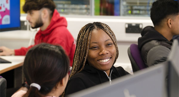 A female is smiling and sitting at a computer desk in the networking lab with peers