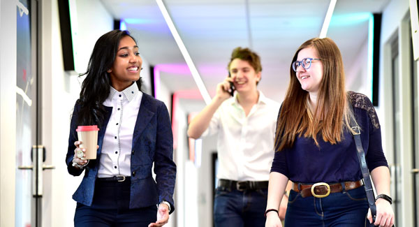 Two females dressed in smart casual clothing are walking and chatting in a corridor, there is a male in the background talking on a mobile phone