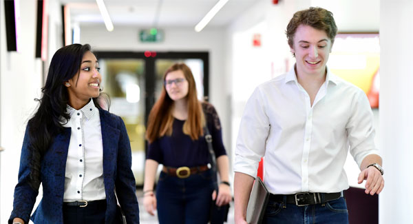 A female and male dressed in smart casual clothing are carrying laptops and chatting while walking in a corridor