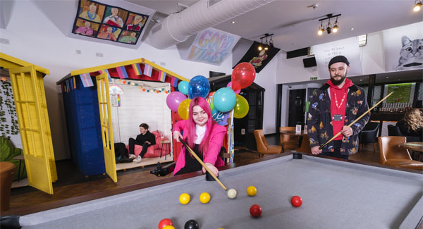 A male and female casually dressed are playing a game of pool at the pub venue on campus, there are other students in the background socialising