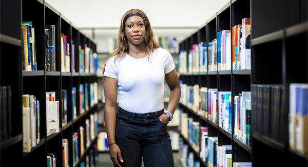 A female student wearing a white t-shirt is standing in the middle of two book shelves in a modern library