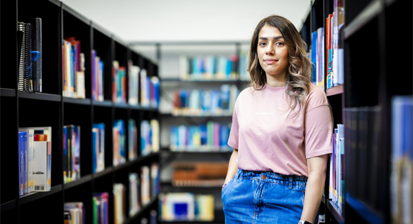 A female student in casual clothing is standing in front of a bookshelf in a modern university library