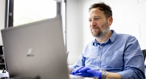 A male with a beard wearing a blue shirt is using a laptop