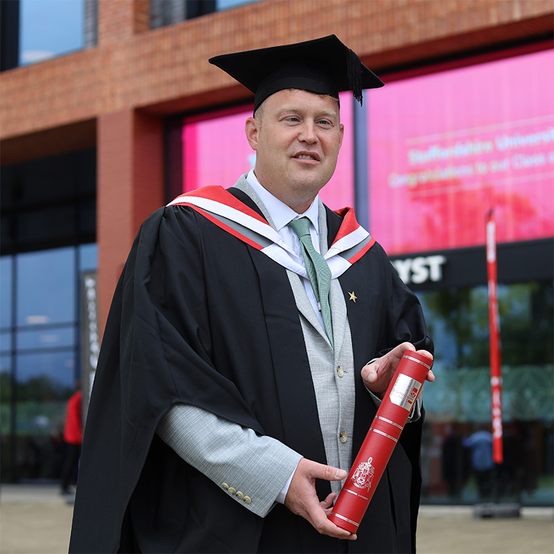 Rob Henshall in his graduation cap and gown holding a scroll