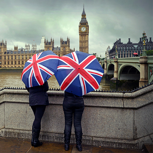 Two people standing underneath umbrellas featuring a Union Jack print looking out to the Palace of Westminster