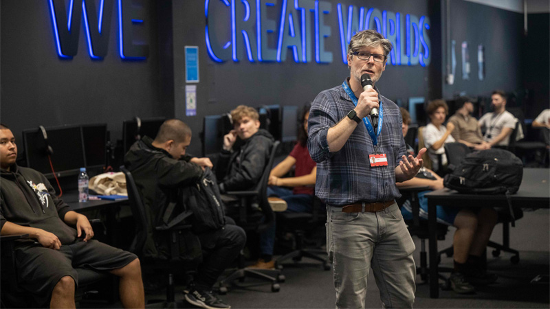 A lecturer is stood up delivering a subject talk to an audience of prospective students in a modern computer lab