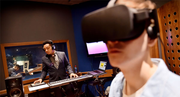 A male wearing a suit and headphones is playing electronic music instruments, behind him is a male working in the music studio