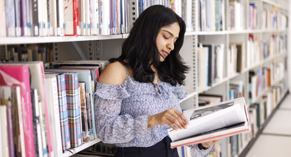 A female with dark hair and floral clothing is standing up reading a book in the University library