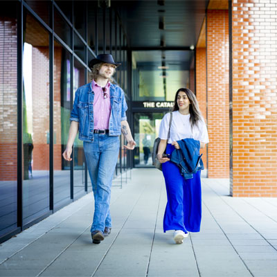 A male and female student wearing blue are chatting whilst walking near the entrance of the Catalyst