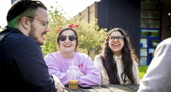 Casually dressed students are chatting and sitting at an outside table on campus