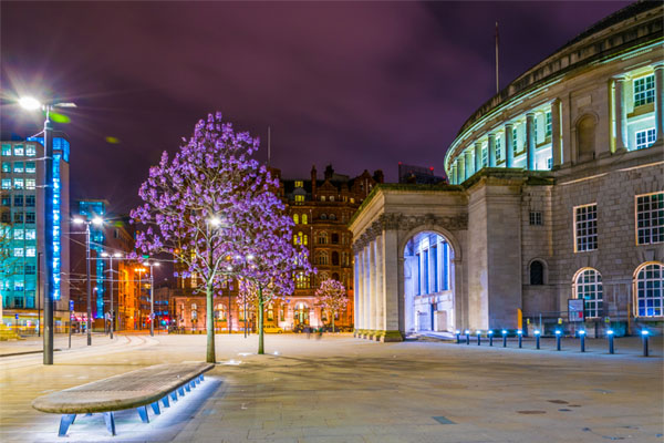 A night time shot of Manchester's historic library lit up, with cherry blossom trees outside and heritage buildings in the distance