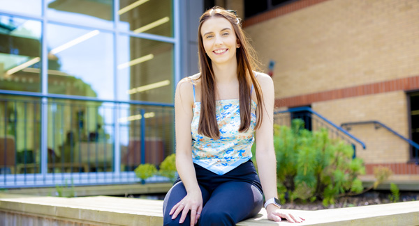 A female with long brown hair is sitting outside the Centre for Health Innovation building on a bright day